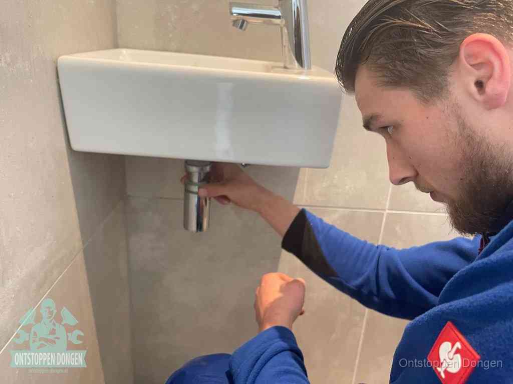Een monteur draait met de hand de zwanenhalssifon onder een fonteinwasbak in de toiletruimte los om de bron van rioollucht in Dongen te vinden