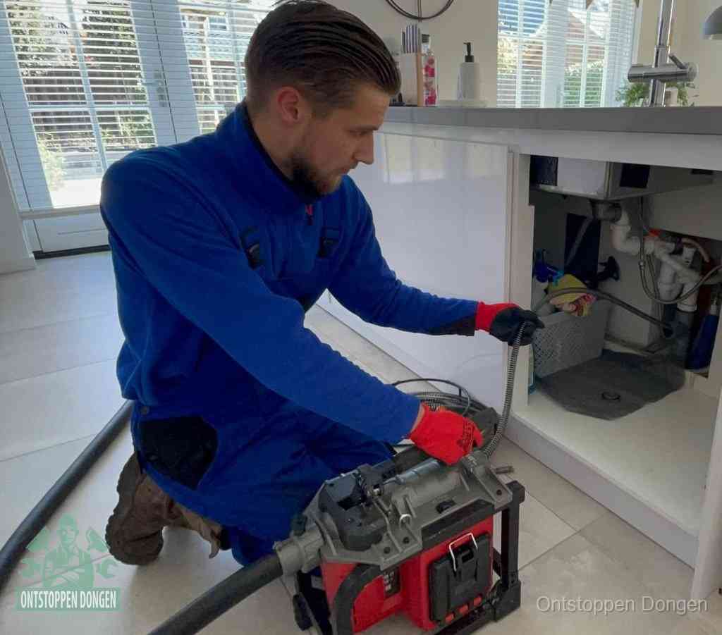 Een monteur met rode handschoenen knielt in een keuken en voert een elektrische ontstoppingsveer in tijdens het professioneel afvoer ontstoppen in Dongen